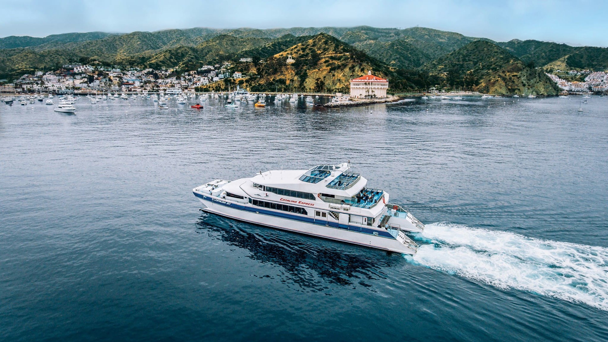 A vibrant dog park bathed in sunlight features abundant greenery with mature trees, offering ample shade for leisurely outdoor activities. In the distance is a well-equipped playground that presents an additional activity area. An inviting picnic table spreads out nearby, ideal for enjoying meals al fresco or taking relaxing breaks from play. A neatly-placed trash bin aids in maintaining the park's cleanliness for everyone's enjoyment. Dominating the scene is a generously wide dirt path gracefully curving through the expanse of grass, promising exploratory walks and lively fetch games.