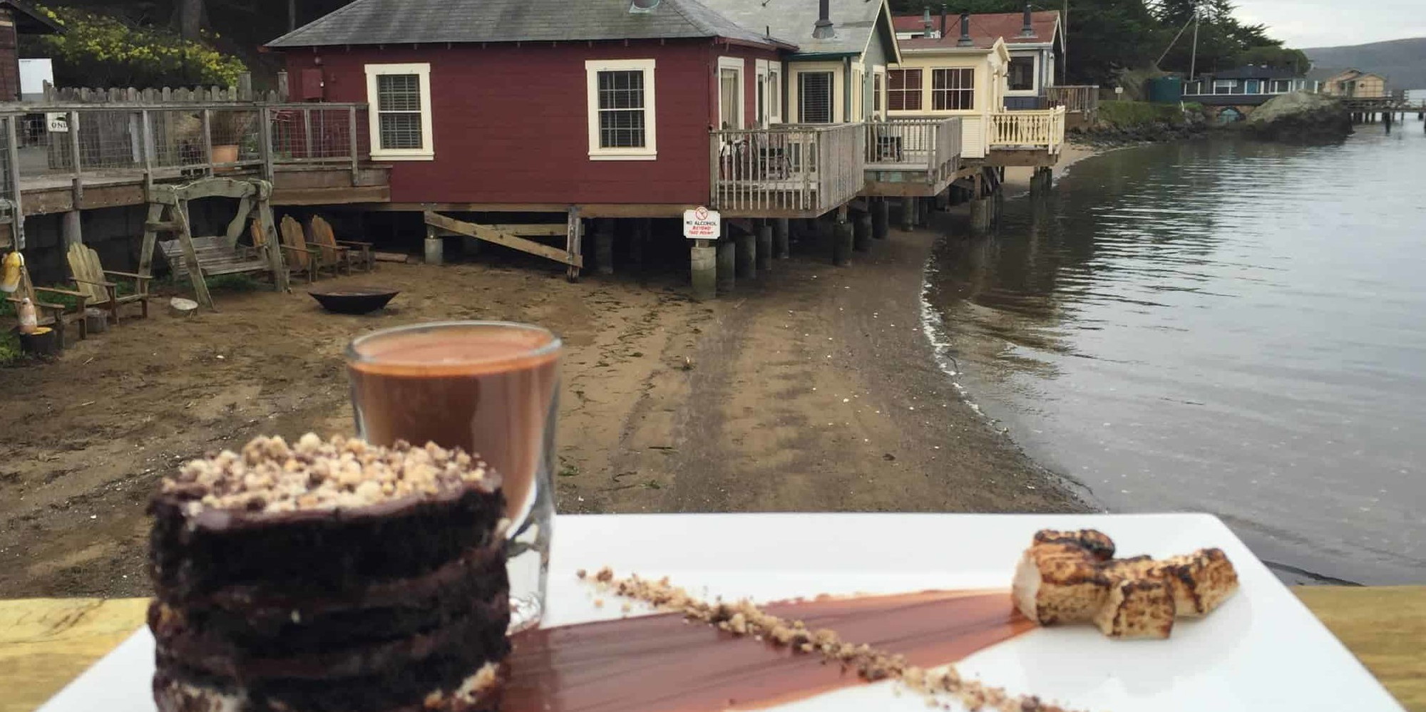 A vibrant dog park bathed in sunlight features abundant greenery with mature trees, offering ample shade for leisurely outdoor activities. In the distance is a well-equipped playground that presents an additional activity area. An inviting picnic table spreads out nearby, ideal for enjoying meals al fresco or taking relaxing breaks from play. A neatly-placed trash bin aids in maintaining the park's cleanliness for everyone's enjoyment. Dominating the scene is a generously wide dirt path gracefully curving through the expanse of grass, promising exploratory walks and lively fetch games.