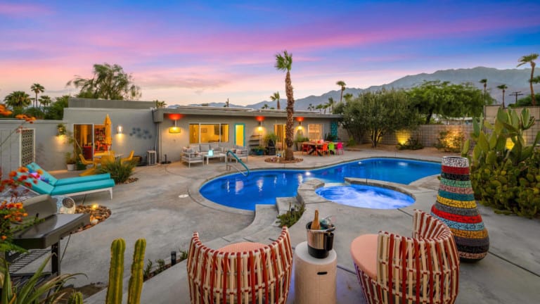 A vibrant dog park bathed in sunlight features abundant greenery with mature trees, offering ample shade for leisurely outdoor activities. In the distance is a well-equipped playground that presents an additional activity area. An inviting picnic table spreads out nearby, ideal for enjoying meals al fresco or taking relaxing breaks from play. A neatly-placed trash bin aids in maintaining the park's cleanliness for everyone's enjoyment. Dominating the scene is a generously wide dirt path gracefully curving through the expanse of grass, promising exploratory walks and lively fetch games.