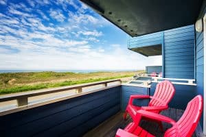 A vibrant dog park bathed in sunlight features abundant greenery with mature trees, offering ample shade for leisurely outdoor activities. In the distance is a well-equipped playground that presents an additional activity area. An inviting picnic table spreads out nearby, ideal for enjoying meals al fresco or taking relaxing breaks from play. A neatly-placed trash bin aids in maintaining the park's cleanliness for everyone's enjoyment. Dominating the scene is a generously wide dirt path gracefully curving through the expanse of grass, promising exploratory walks and lively fetch games.