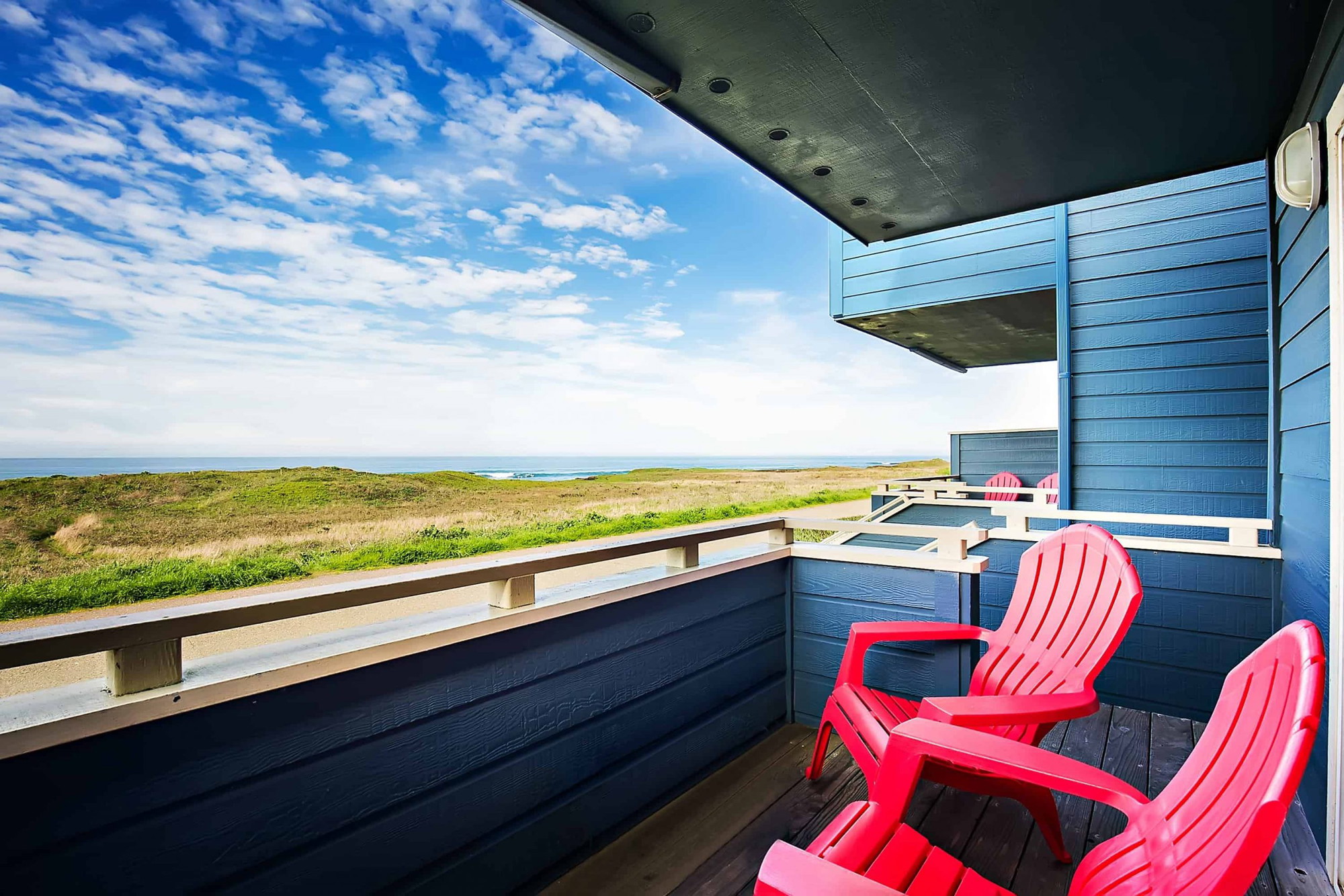 A vibrant dog park bathed in sunlight features abundant greenery with mature trees, offering ample shade for leisurely outdoor activities. In the distance is a well-equipped playground that presents an additional activity area. An inviting picnic table spreads out nearby, ideal for enjoying meals al fresco or taking relaxing breaks from play. A neatly-placed trash bin aids in maintaining the park's cleanliness for everyone's enjoyment. Dominating the scene is a generously wide dirt path gracefully curving through the expanse of grass, promising exploratory walks and lively fetch games.