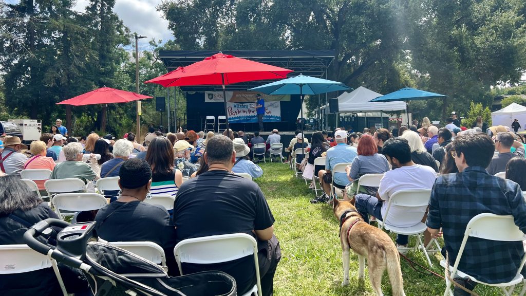 A crowd of people sits on white chairs arranged outdoors, all facing a stage with a banner titled 